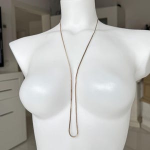Puede incluir: Un delicado collar de color dorado exhibido en un maniquí blanco. El collar presenta una fina cadena plana que cae por la parte delantera, creando un aspecto simple pero elegante. El maniquí está en una habitación bien iluminada.