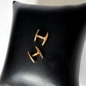May include: A pair of gold-colored cufflinks, each with a distinctive T-shape design, rest on a black leather-like surface. The cufflinks are positioned diagonally, showcasing their polished finish and intricate details. The background is a solid black, providing a stark contrast.