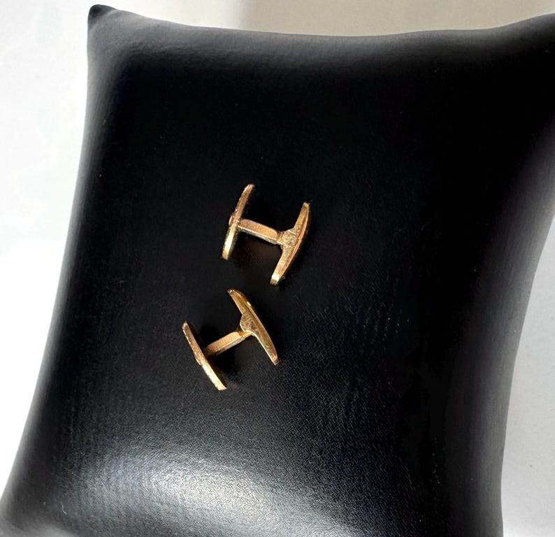 May include: A pair of gold-colored cufflinks, each with a distinctive T-shape design, rest on a black leather-like surface. The cufflinks are positioned diagonally, showcasing their polished finish and intricate details. The background is a solid black, providing a stark contrast.