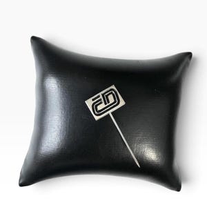 May include: A silver-toned lapel pin featuring a rectangular head with a black, stylized geometric logo design. The pin has a long, thin, straight metal needle extending from the bottom of the rectangular head, designed for attachment to fabric.