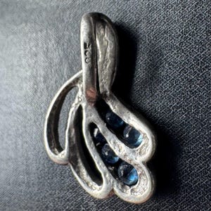 Op de afbeelding: Zilveren hanger met een delicaat, abstract ontwerp met drie rijen blauwe edelstenen. De hanger heeft een lus aan de bovenkant om een ​​ketting aan te bevestigen. De achterkant van de hanger is gestempeld met het nummer "925", wat aangeeft dat hij van sterling zilver is.