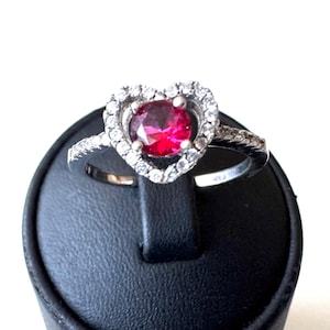 May include: A silver ring with a heart-shaped design. The center features a vibrant red gemstone, surrounded by a halo of smaller, clear stones. The band is also adorned with clear stones. The ring is displayed on a black stand.