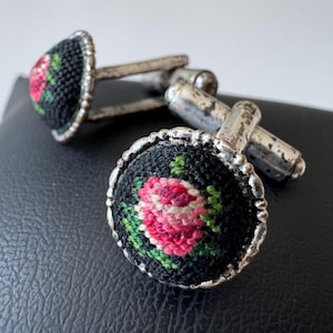 Könnte beinhalten: Ein Paar Vintage-Manschettenknöpfe. Jeder Manschettenknopf hat einen schwarzen Hintergrund mit einem gestickten Design einer rosa Rose mit grünen Blättern. Sie sind von einem silberfarbenen Metallrand und -pfosten umgeben.