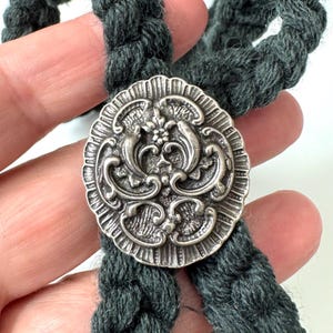 May include: A dark gray, braided rope necklace with a decorative silver-toned button clasp. The button features an ornate, floral design. The rope is thick and appears to be made of a soft, woven material.