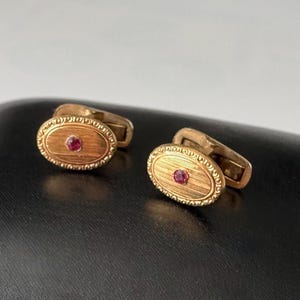 Könnte beinhalten: Ein Paar goldfarbene Manschettenknöpfe mit ovalem Design. Jeder Manschettenknopf hat einen kleinen, runden, roten Edelstein in der Mitte. Die Manschettenknöpfe haben eine strukturierte Oberfläche und einen klassischen, eleganten Stil.