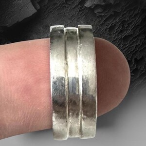 Può includere: Un ampio anello in argento caratterizzato da un design a tre fasce con due distinte scanalature orizzontali che attraversano il centro della superficie metallica lucida, creando un aspetto a strati sulla fascia che avvolge il dito.
