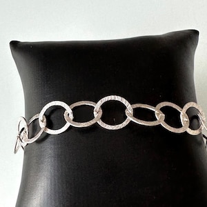 May include: A silver chain bracelet with textured oval links. The bracelet is displayed on a black pillow.