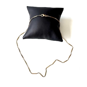 May include: A gold-coloured necklace with a delicate design, featuring small, elongated beads and a secure clasp. The necklace is displayed on a black cushion against a white background, showcasing its elegant simplicity.