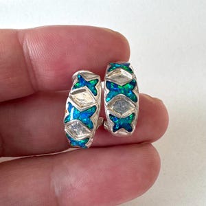 Puede incluir: Un par de pendientes de aro plateados con un diseño geométrico. Cada pendiente presenta un patrón de incrustaciones de ópalo azul y verde y detalles plateados. Los pendientes son pequeños y parecen ligeros.