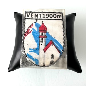 Könnte beinhalten: Ein weißer Stoff-Patch mit schwarzen Nähten und dem Text "VENT 1900m". Der Patch zeigt eine bunte Illustration einer Kirche mit einem hohen Kirchturm und einem Berg im Hintergrund.