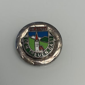 Puede incluir: Un pin de color plateado con un diseño de esmalte verde, azul y rojo. El pin presenta una iglesia con un techo rojo y una colina verde en el fondo. El texto "DEM TREUEN GAST" está escrito alrededor de la iglesia en un círculo. El texto "FVL. HIPPACH" está escrito encima de la iglesia.