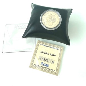 Könnte beinhalten: Eine Goldmünze mit Echtheitszertifikat. Die Münze zeigt ein Motiv des deutschen Bundesadlers und den Text "50 Jahre Bundesrepublik Deutschland". Das Zertifikat ist weiß mit schwarzem Text und dem Text "50 Jahre BRD" und "A 0572".