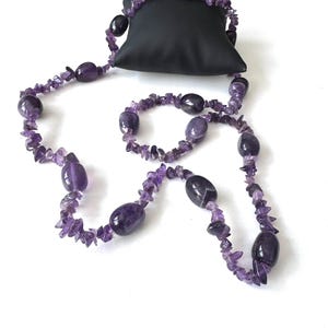 May include: A purple amethyst necklace with a mix of polished oval beads and small, irregular chips. The necklace is draped over a small black pillow, showcasing its color and design. The gemstones are a deep, rich purple.