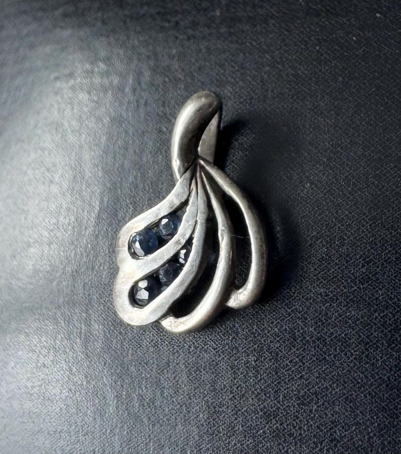 Op de afbeelding: Een zilveren hanger met een gestileerd ontwerp met meerdere gebogen lijnen en kleine blauwe edelstenen.
