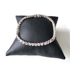 Könnte beinhalten: Ein silberfarbenes Tennisarmband mit einem sich wiederholenden S-förmigen Metallglieddesign. Das Armband ist durchgehend mit abwechselnd klaren und hellrosa rund geschliffenen Steinen besetzt und verfügt über einen Metall-Kastenschlossverschluss.