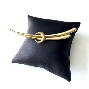 May include: A gold-toned brooch with a curved, abstract design. The brooch features a circular element at the center, with two curved bars extending outwards. The brooch is displayed on a black pillow against a white background.