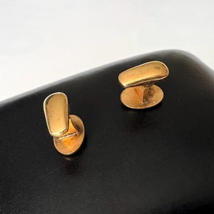 May include: A pair of gold-colored cufflinks. Each cufflink features a rectangular top and an oval base, connected by a small post. The cufflinks are displayed on a black surface.