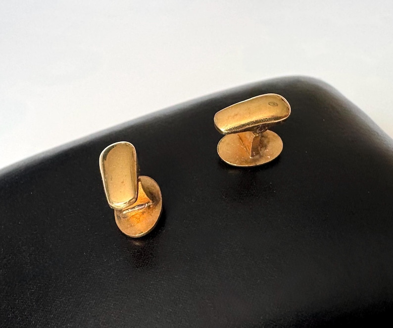 May include: A pair of gold-colored cufflinks. Each cufflink features a rectangular top and an oval base, connected by a small post. The cufflinks are displayed on a black surface.