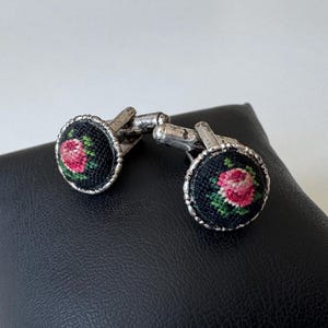 Könnte beinhalten: Ein Paar Vintage-Manschettenknöpfe mit floralem Design. Die runden, silberfarbenen Manschettenknöpfe haben einen schwarzen Hintergrund mit einer detaillierten gestickten Rose in Rosa- und Grüntönen. Die Manschettenknöpfe werden auf einer schwarzen Oberfläche präsentiert.