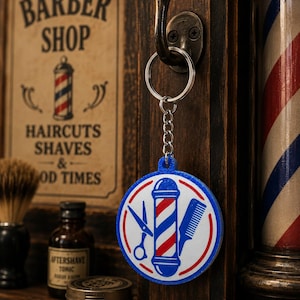 May include: A circular keychain featuring a graphic of a red, white, and blue barber pole flanked by a pair of scissors and a comb. The design is enclosed in a blue circular border with a silver metal ring and chain attached to the top loop.