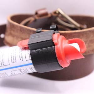 May include: A black plastic holster with a red pepper spray canister attached to a brown leather belt. The holster has a clip to attach to a belt.