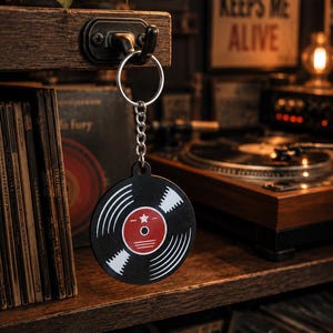 Pode incluir: Um porta-chaves circular com um gráfico de disco de vinil preto e branco com um rótulo central vermelho. O design inclui sulcos brancos e uma pequena estrela branca no rótulo vermelho. O porta-chaves está preso a um anel e corrente de metal prateado.