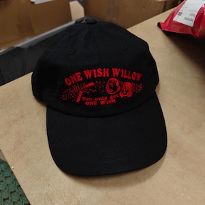 May include: A black baseball cap featuring red embroidered text that reads ONE WISH WILLOW and YOU ONLY GET ONE WISH, accompanied by a small red graphic illustration of two faces positioned between the lines of text on the front panel.