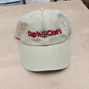 Peut inclure: Une casquette de baseball beige avec une construction à six panneaux et une visière incurvée. L'avant présente un texte rouge brodé indiquant Cap'n Clark's, avec une petite icône de roue de gouvernail marron remplaçant l'apostrophe.