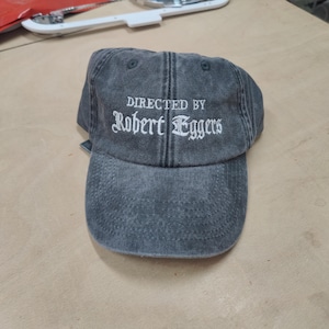Può includere: Un cappello da baseball grigio scuro con testo ricamato bianco che recita "DIRECTED BY Robert Eggers".