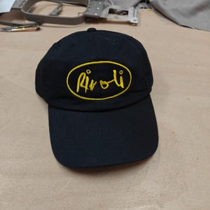 Op de afbeelding: Zwarte baseballpet met een gebogen klep. De pet heeft een gele, geborduurde ovale patch met het woord "Rivoli" in een gestileerd lettertype. De pet is gemaakt van een duurzame stof en heeft een klassieke, casual stijl.