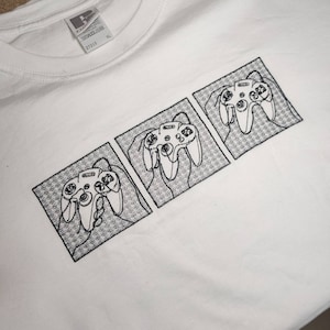 Può includere: Maglietta bianca con un design ricamato nero di tre controller Nintendo 64 in fila.