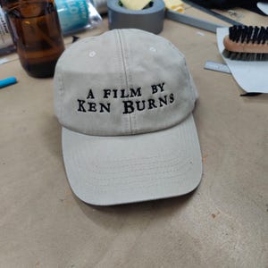 May include: A beige baseball cap with black embroidered text that reads "A Film By Ken Burns".