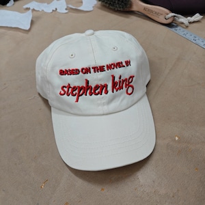 Könnte beinhalten: Cremefarbene Baseballkappe mit dem gestickten Text "BASED ON THE NOVEL BY" über "stephen king" in Rot. Die Kappe hat einen gebogenen Schirm und einen Knopf oben. Ein klassisches Accessoire für Stephen King-Fans.