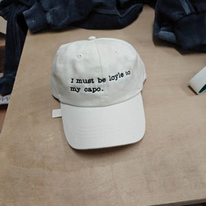 May include: Cream-coloured baseball cap with a curved peak. The cap has the embroidered text "I must be loyle to my capo" in black. The hat is made of a soft fabric and has a small adjustable strap.
