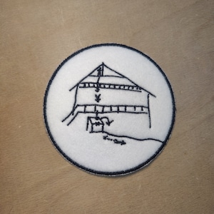 May include: A round white patch featuring a black embroidered illustration of a building with arrows and lines. The patch has a black border, set against a light brown surface.
