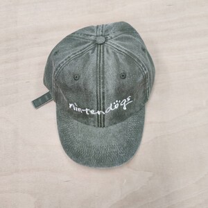 K&ouml;nnte beinhalten: Eine gr&uuml;ne, abgenutzte Baseballkappe aus Baumwolle mit einem wei&szlig;en gestickten Logo, auf dem "nintendogs" steht.