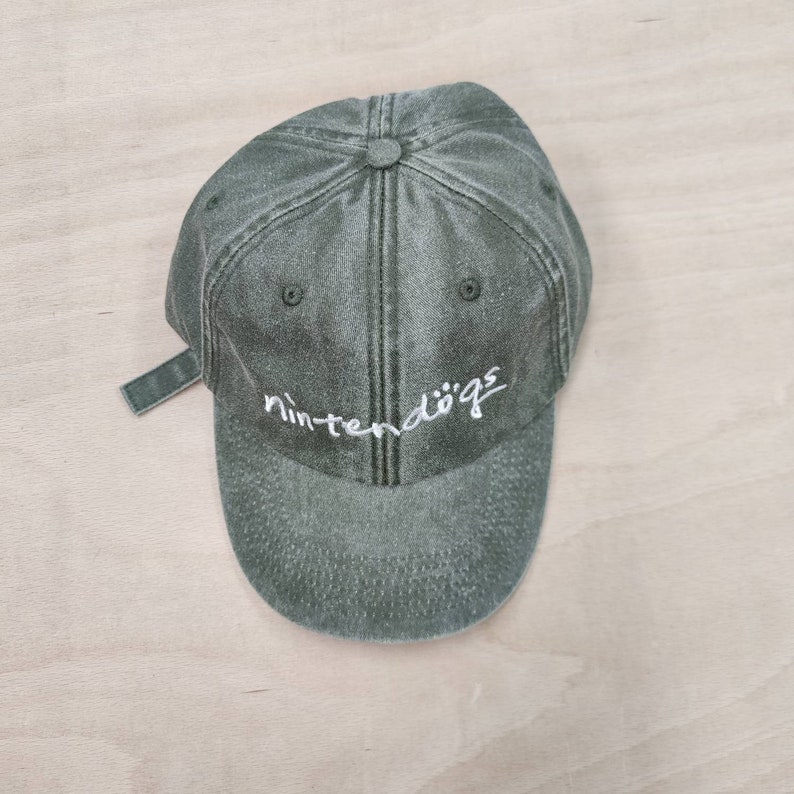 K&ouml;nnte beinhalten: Eine gr&uuml;ne, abgenutzte Baseballkappe aus Baumwolle mit einem wei&szlig;en gestickten Logo, auf dem "nintendogs" steht.