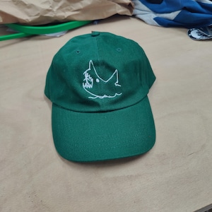 Op de afbeelding: Een donkergroene baseballcap met een wit geborduurd haaienontwerp. De cap heeft een gebogen klep en een stoffen bandsluiting. Het haaienontwerp is een eenvoudige omtrek van een haaienkop met gekartelde tanden.