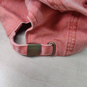 May include: Close-up of a coral-coloured baseball cap with a brass buckle on the back strap.