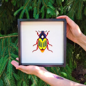 May include: A framed paper art piece of a colourful beetle with red, yellow, and blue accents. The beetle is displayed on a white background.