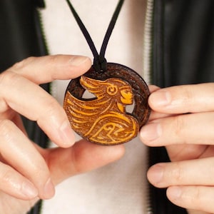 May include: A round, wooden pendant necklace featuring a carved design of a woman's profile with flowing hair. The pendant is brown and tan, suspended from a black cord. The necklace is held against a black leather jacket.
