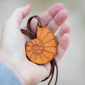 May include: Wooden pendant necklace featuring a carved nautilus shell design. The pendant is a warm orange color with a brown leather cord. The nautilus shell design is a spiral shape.