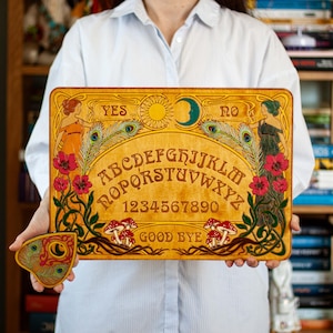 May include: A wooden Ouija board with a matching planchette. The board features an alphabet, numbers, and the words "YES" and "NO". Decorative elements include illustrations of women, flowers, peacock feathers, a sun, and a crescent moon. The words "GOOD BYE" are also present.