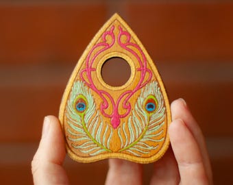 May include: Wooden planchette with a decorative design. The planchette features a central hole and is adorned with pink and teal peacock feather accents. The planchette is held in a person's hand against a blurred brick background.