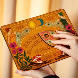 Op de afbeelding: Een houten Ouija-bord met illustraties in Art Nouveau-stijl. Het bord toont het alfabet, cijfers en de woorden "YES", "NO" en "GOOD BYE". Het is versierd met pauwenveren, bloemen en figuren in oranje en groen.