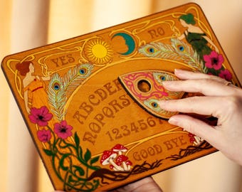 Könnte beinhalten: Ein hölzernes Ouija-Brett mit Illustrationen im Jugendstil. Das Brett zeigt das Alphabet, Zahlen und die Wörter "YES", "NO" und "GOOD BYE". Es ist mit Pfauenfedern, Blumen und Figuren in Orange und Grün verziert.