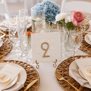 May include: A square white table number sign with an embossed pattern and a gold number 2, displayed on a gold wire stand. The setting includes woven placemats, white plates, gold cutlery, clear glassware, and floral arrangements with blue and pink flowers.