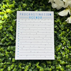 May include: A white notepad with a blue and gray design. The notepad has the words "Procrastination Agenda" printed on the top. The notepad is on a green background.