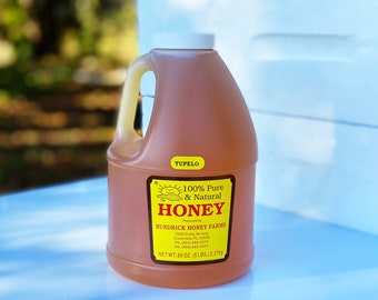 May include: A gallon jug of 100% pure and natural Tupelo honey from Bundrick Honey Farms. The label is yellow with a sun and the words "100% Pure & Natural Honey" in red. The jug is sitting on a white surface.