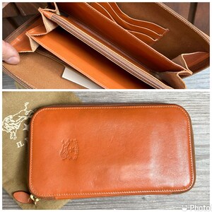 May include: A brown leather zippered wallet with the brand name "IL Bisonte" embossed on the front. The wallet is closed and lying on a brown fabric surface.